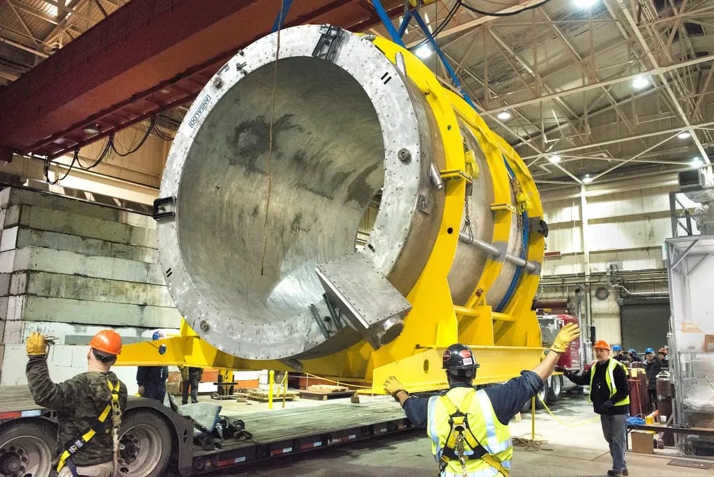 Workers in high-visibility clothing guiding a large cylindrical machine assembly onto a low-loader inside a workshop.