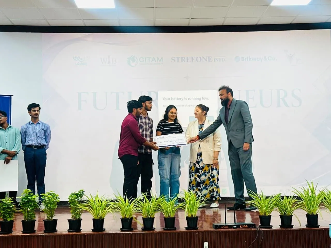 A cheque being presented to student founders on stage at the Futurepreneurs segment, under a banner carrying the partner organisations.