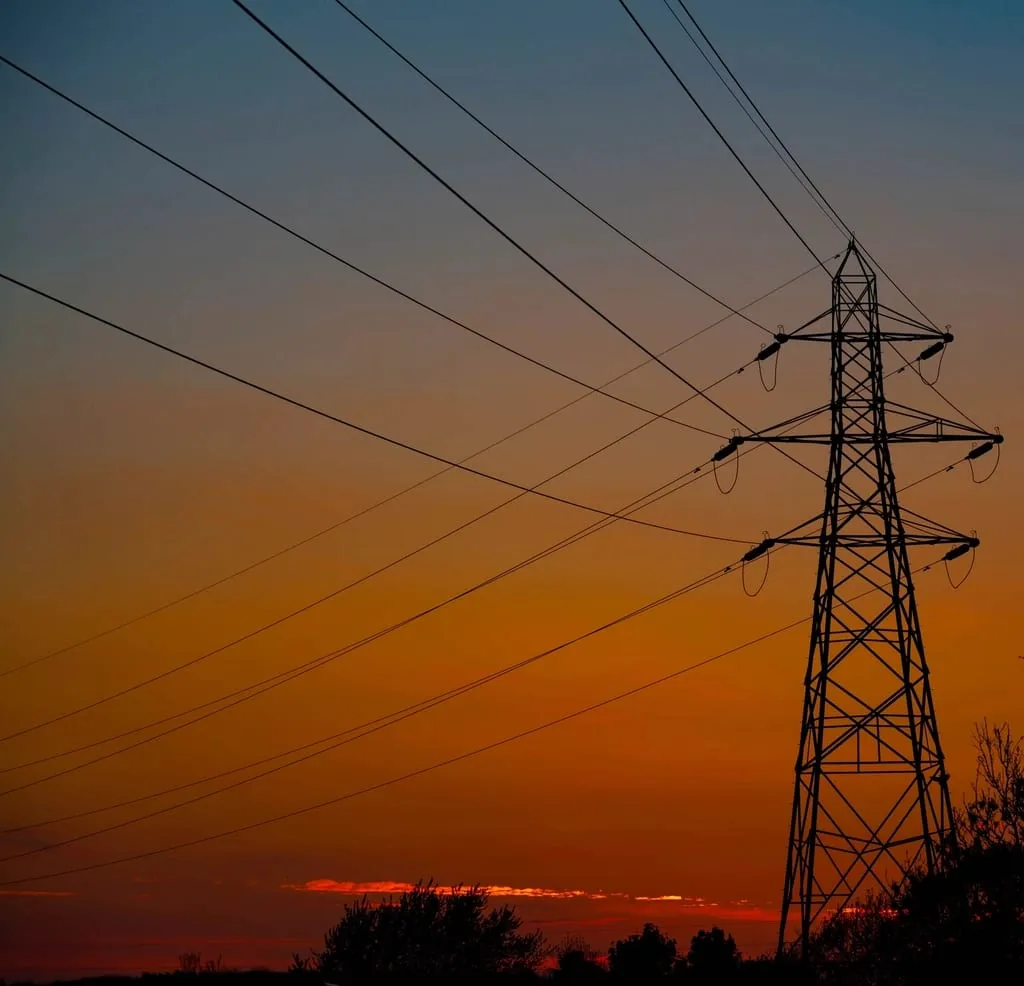 An electricity transmission pylon and conductors silhouetted against a sunset sky.